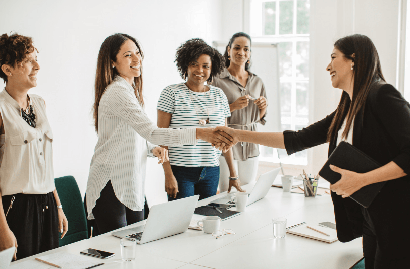A business woman shaking the hands of a business owner who is sitting amongst collegues being interviewed about their marketing strategies