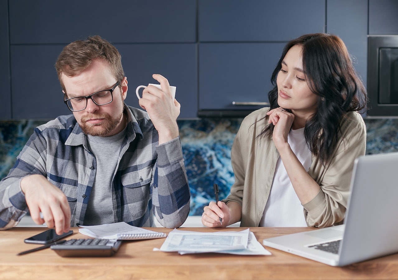 A couple of business partners sitting in their kitchen with a calculator, laptop and coffee, reviewing their finances using Digital Prosperity Podcast's six step system