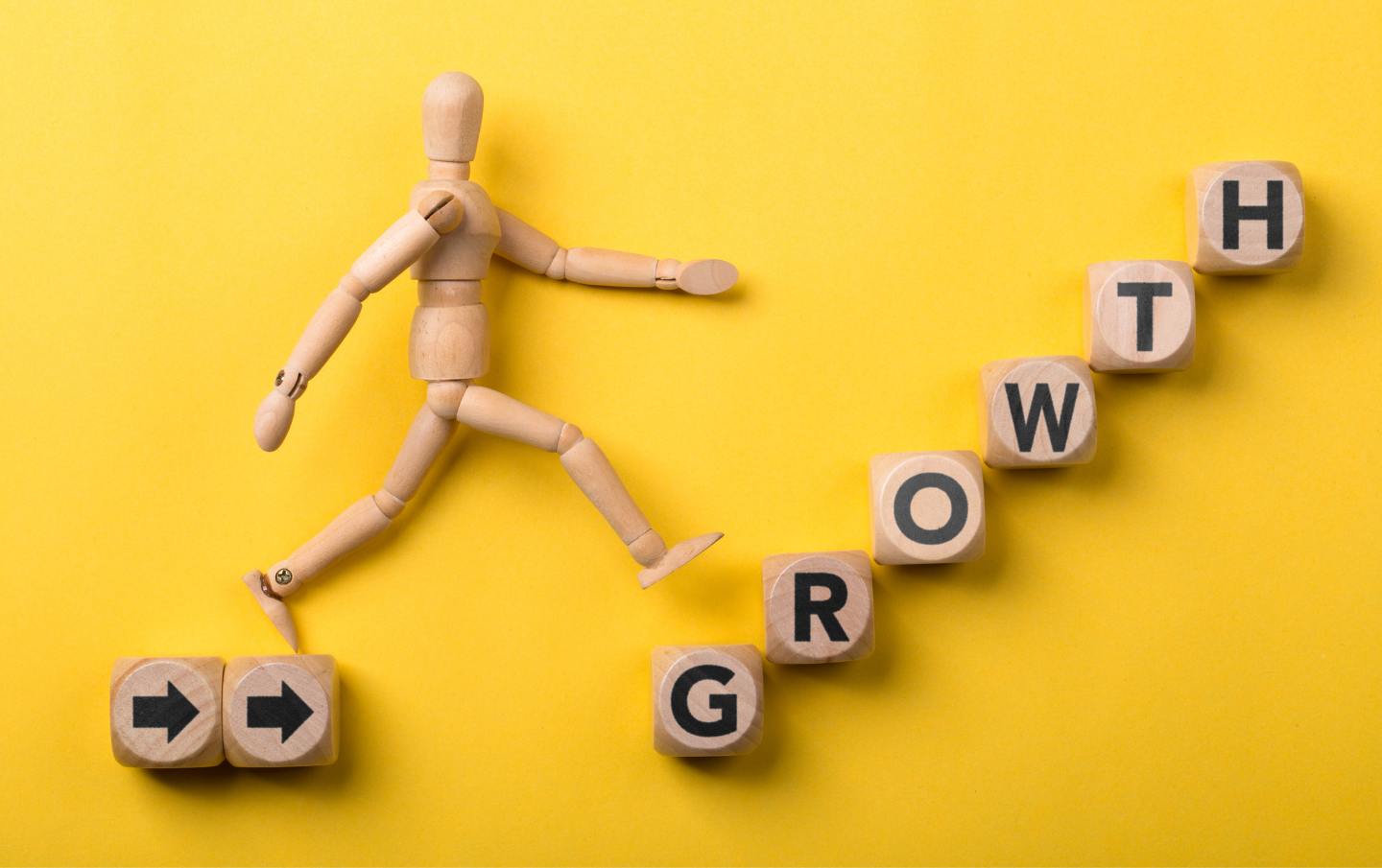 A wooden figure climbing wooden building blocks that spell out the word Growth as the figure's small business grows in profit and size.