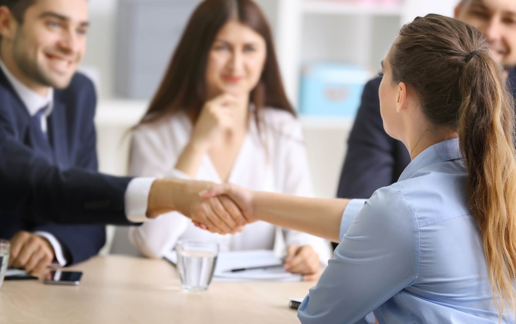 A woman shaking the hands of a business owner as she sits at a table across from him and two other managers about to be interviewed for a position at a company overcoming their recruitment barriers.