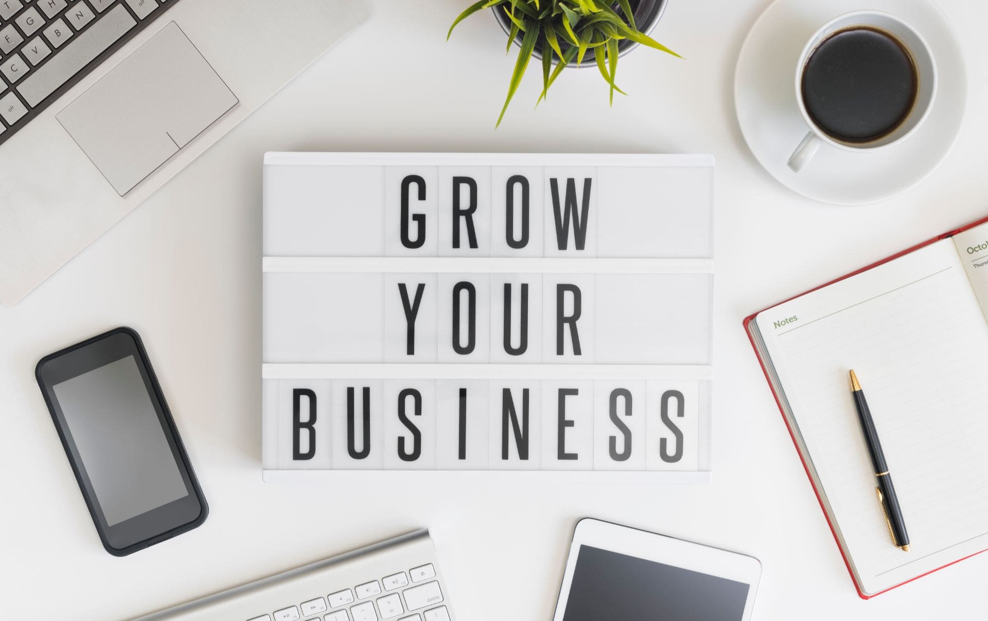 A sign on the desk of a business owner that reads Grow Your Business, with the business owner's mobile, laptop, coffee and diary around it as they use HubSpot sales tools for growth.