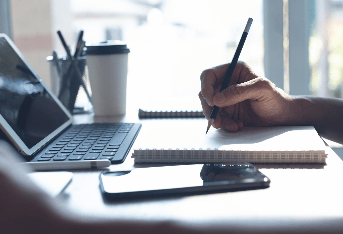 A person making notes on their desk next to a laptop and phone.