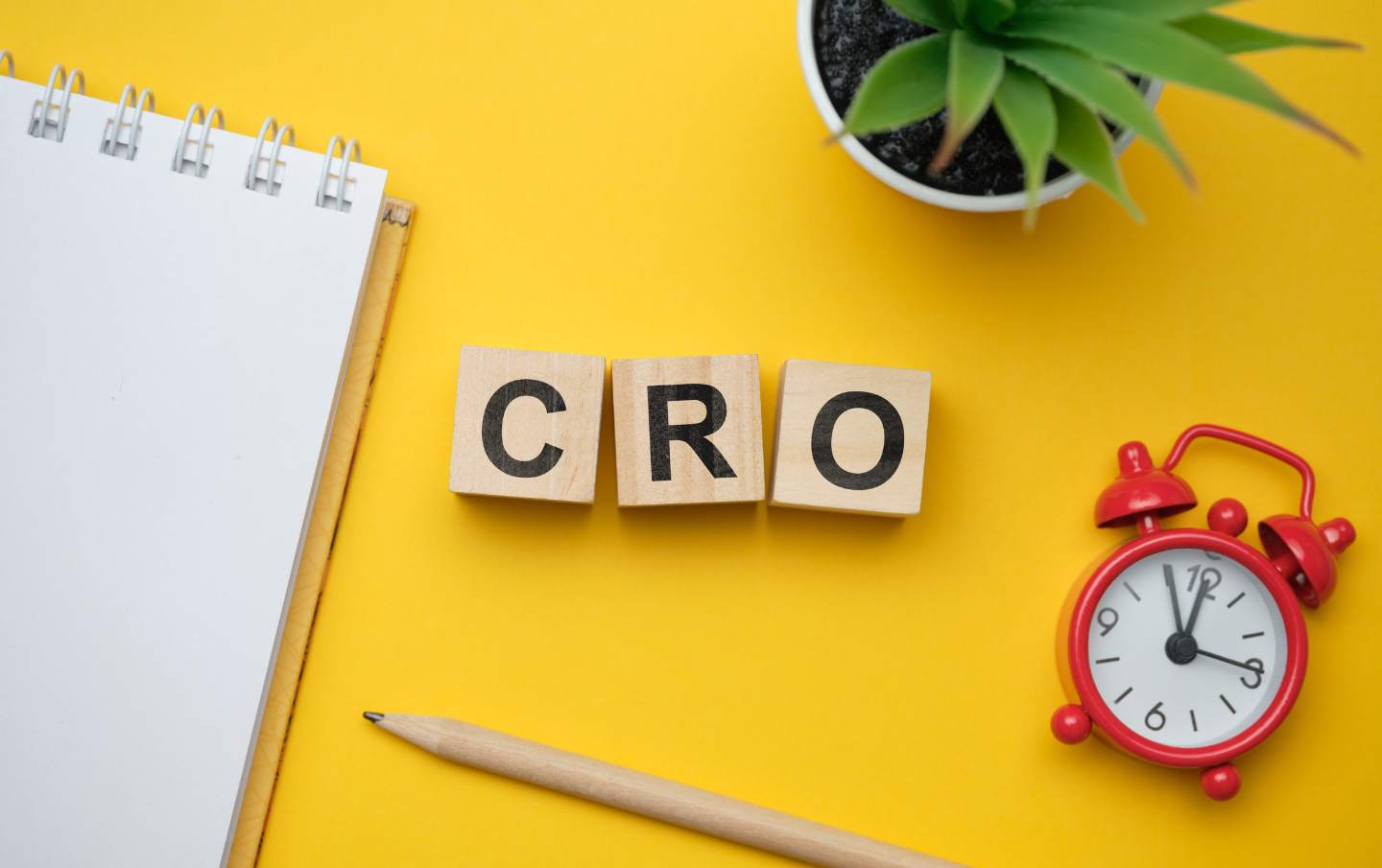 Wooden blocks spelling out C R and O for conversion rate optimisation surrounded by a notepad, pencil, desk plant and alarm clock, on a business owners yellow desk