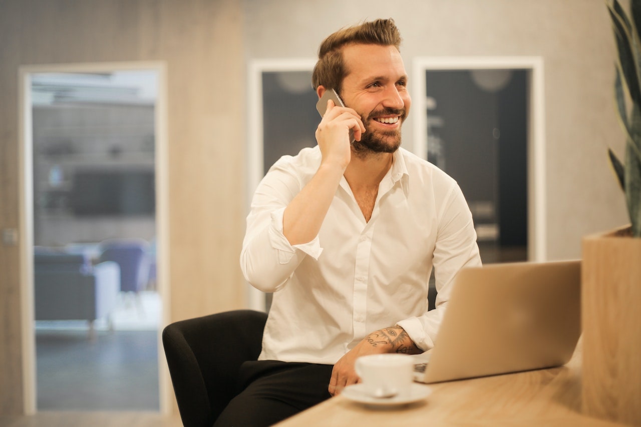A business sales representative sitting at a table with a mug and laptop, calling a prospect to follow up after meeting with them earlier that week. 