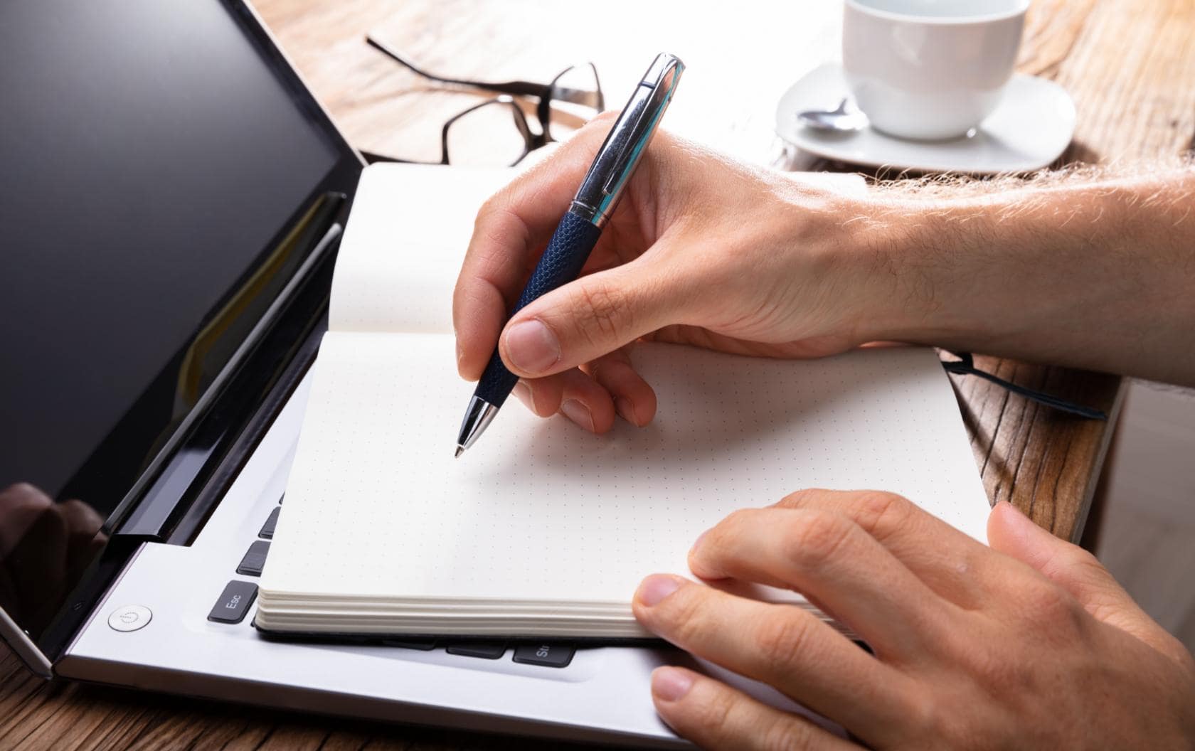 A business owner writing in a notebook placed on top of his laptop keyboard, writing a strategy on how to use content marketing for his business to elevate and grow. 