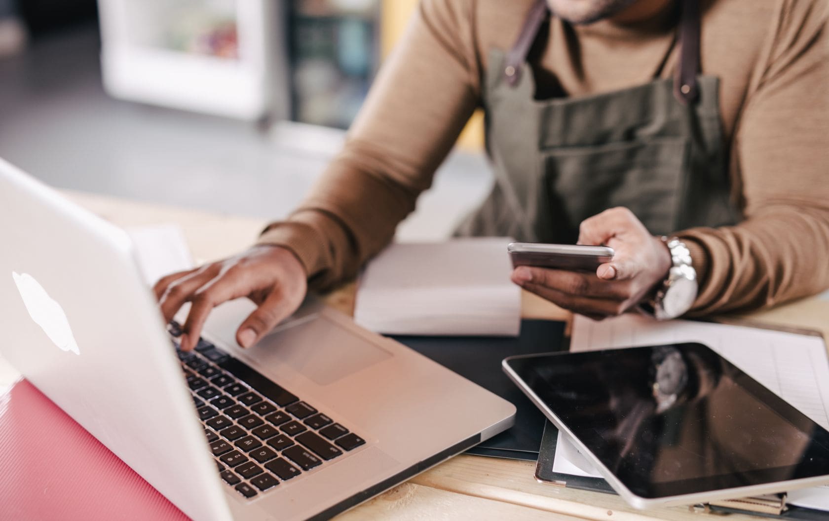 A small business owner in an apron sitting at his desk using HubSpot on his laptop, while making sales on his phone with his iPad on a pile of files.