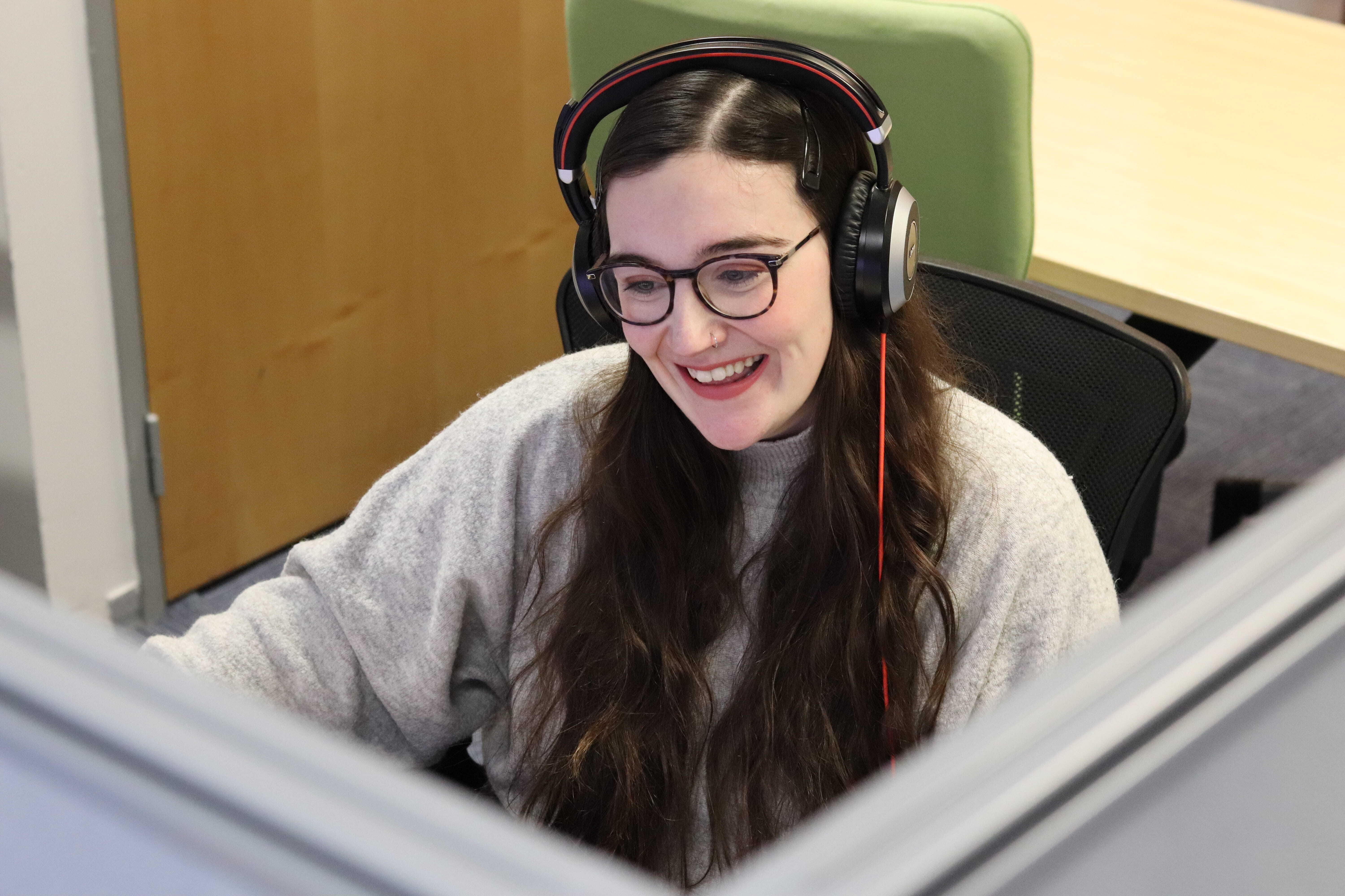 Danni wearing glasses and her hadset, smiling and looking down at her computer.