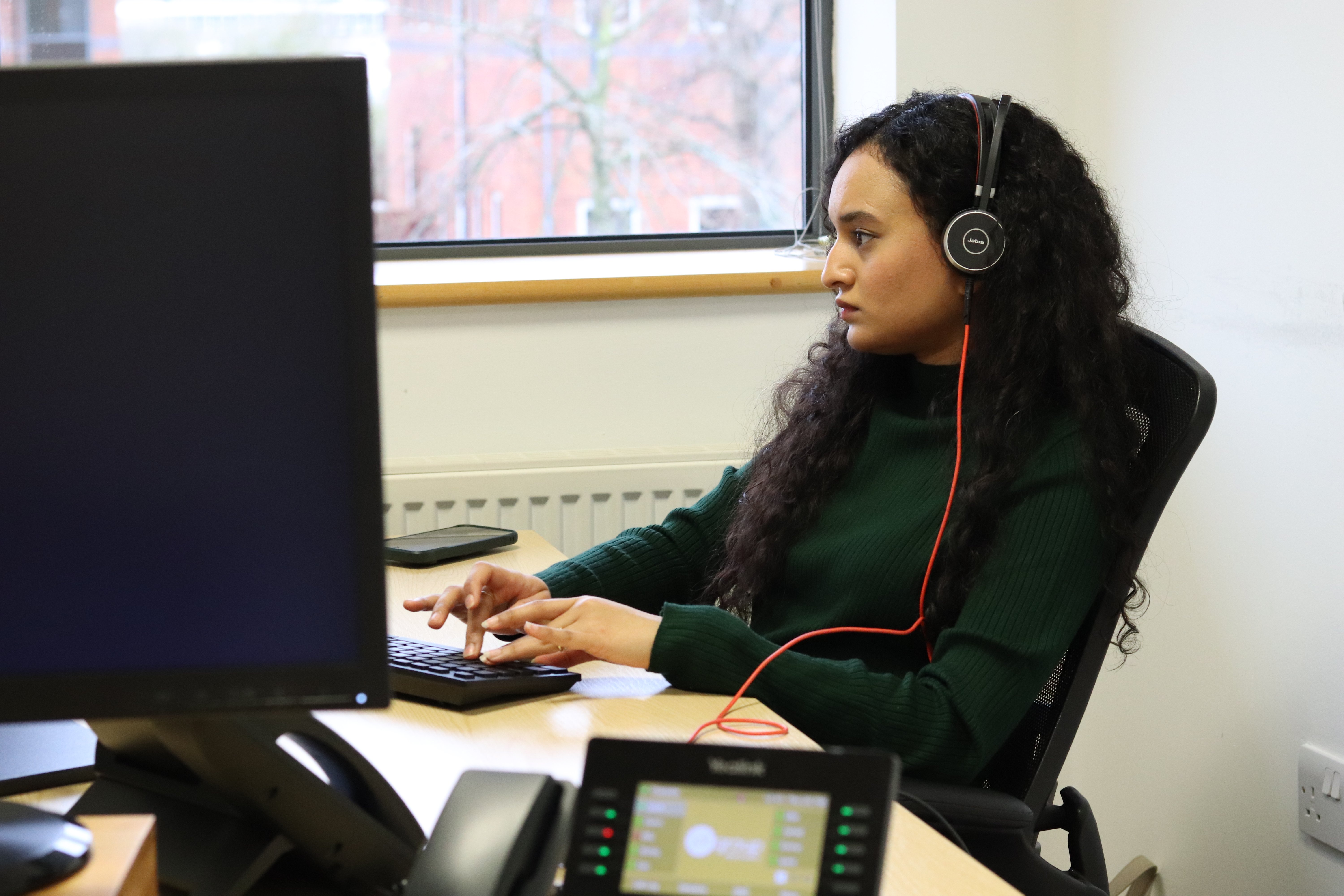 Ash with her headset on typing on her black keyboard while focusing on her computer monitors.