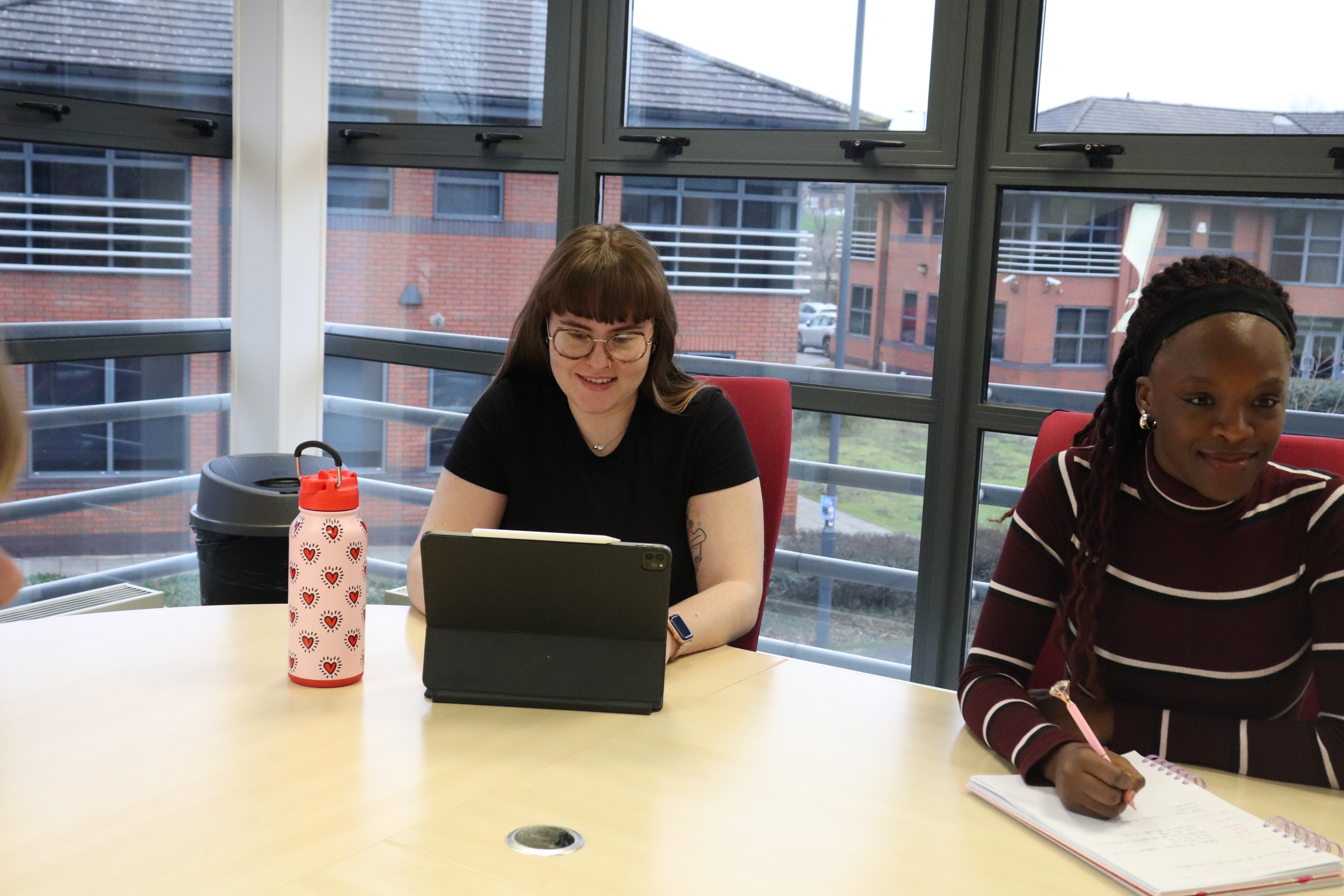 Ellis typing on her I pad and Mia writing in a notebook, in a meeting about marketing strategies for coaches.