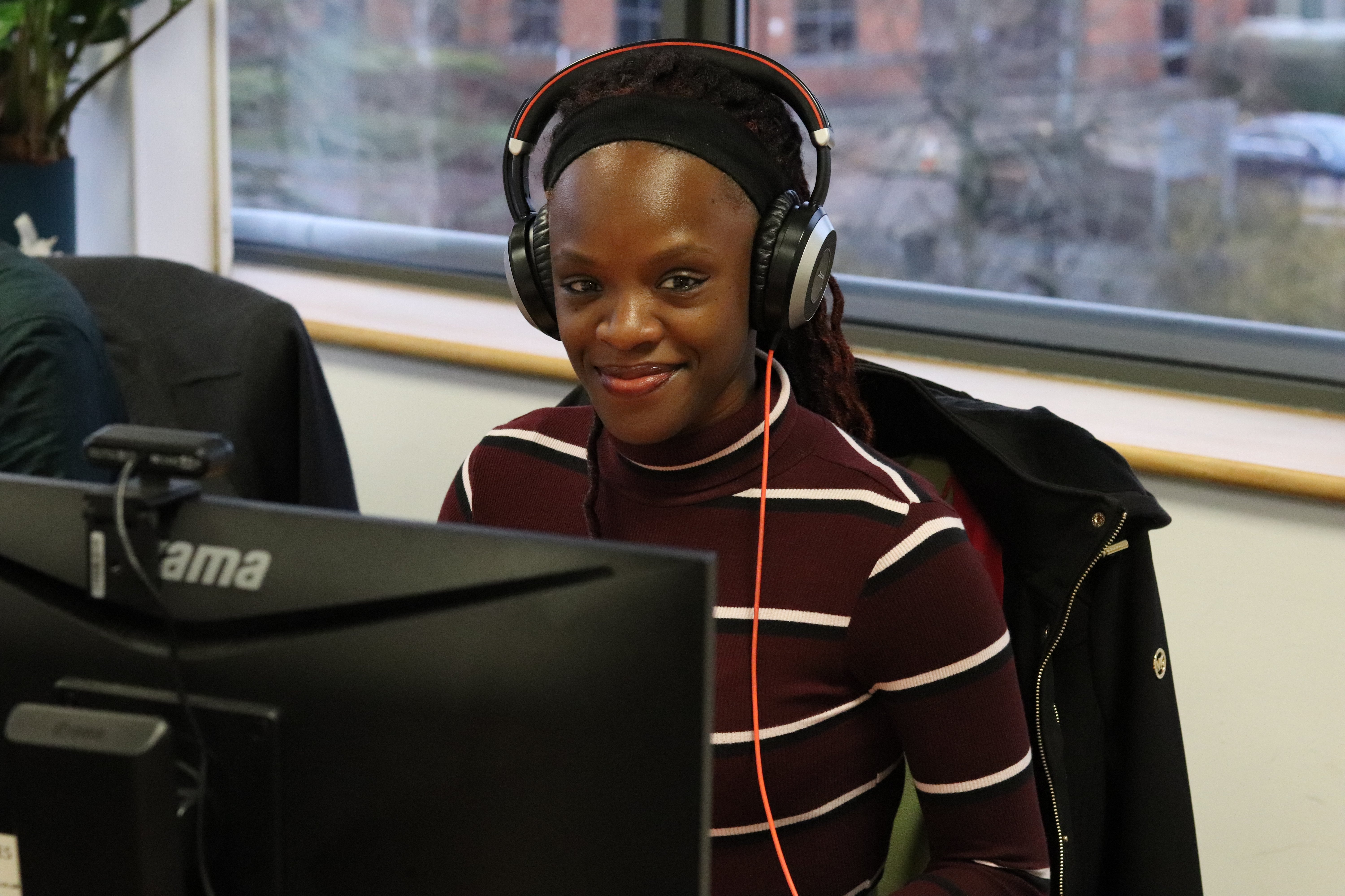 Mia wearing a striped long sleeve t shirt smiling at the camera with her headset on at her desk.
