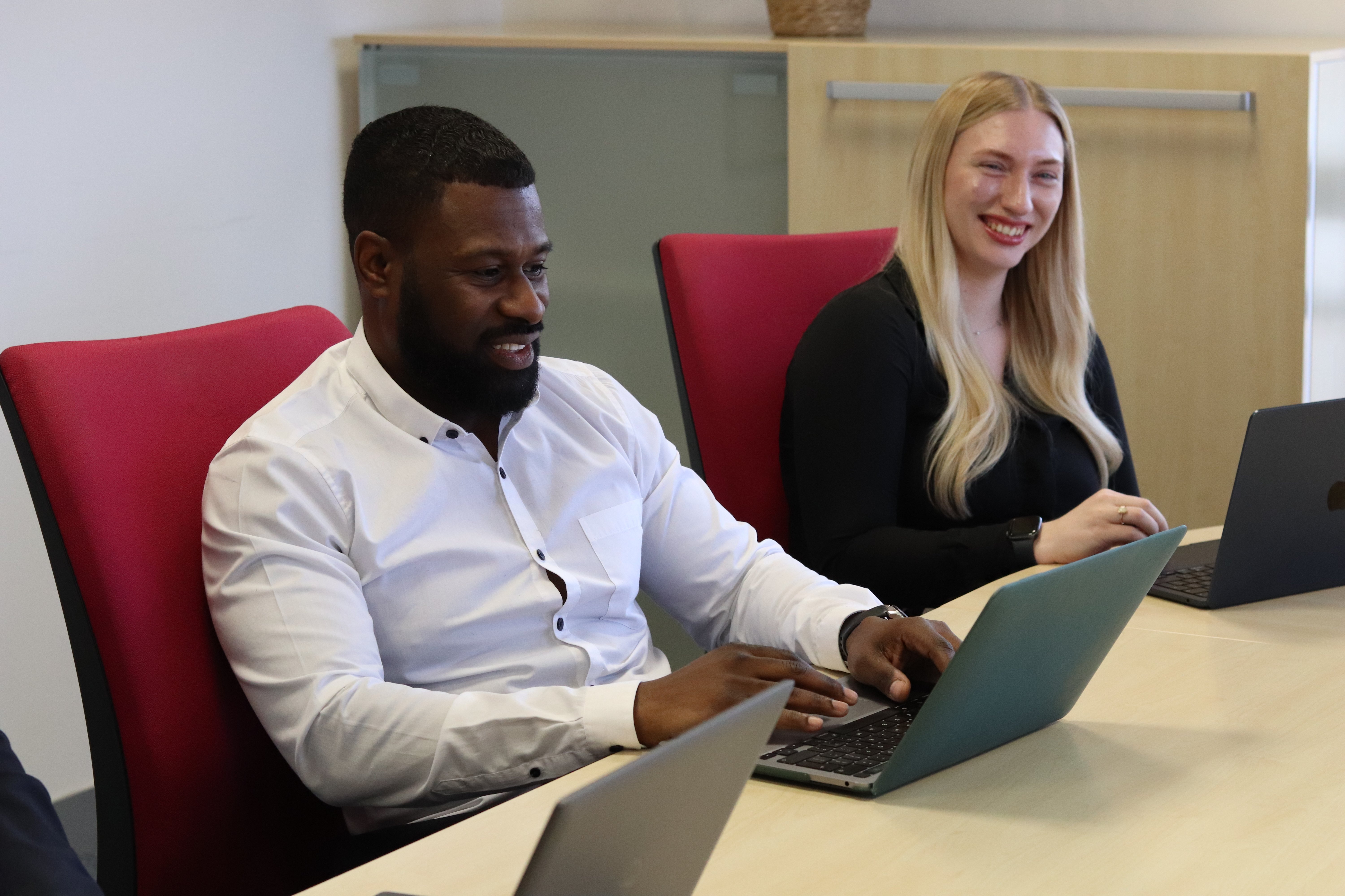 Dan and Amber smiling and talking while on their laptops in a meeting.