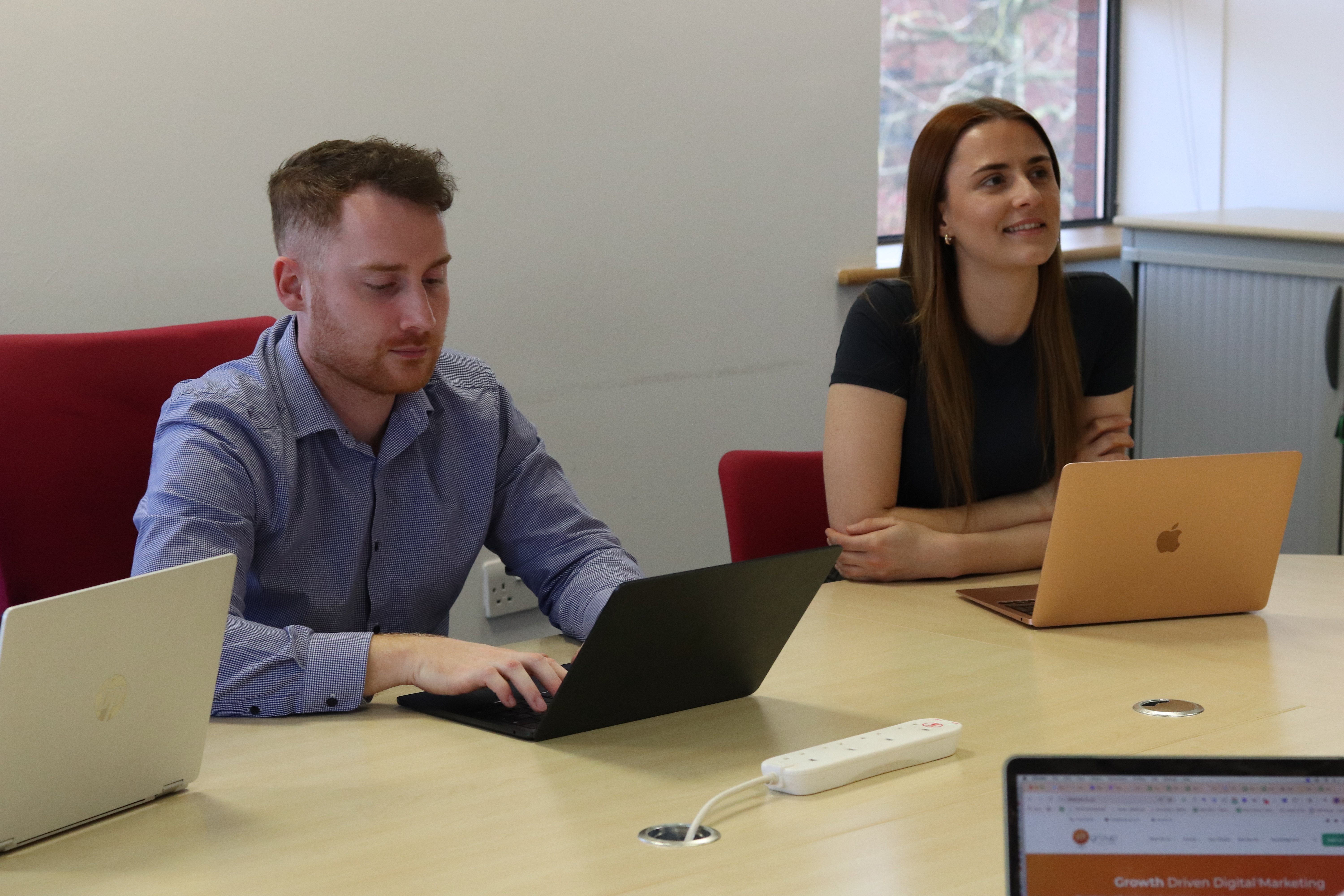 John typing on his laptop and Charlotte sitting next to him looking across at the person leading the meeting.