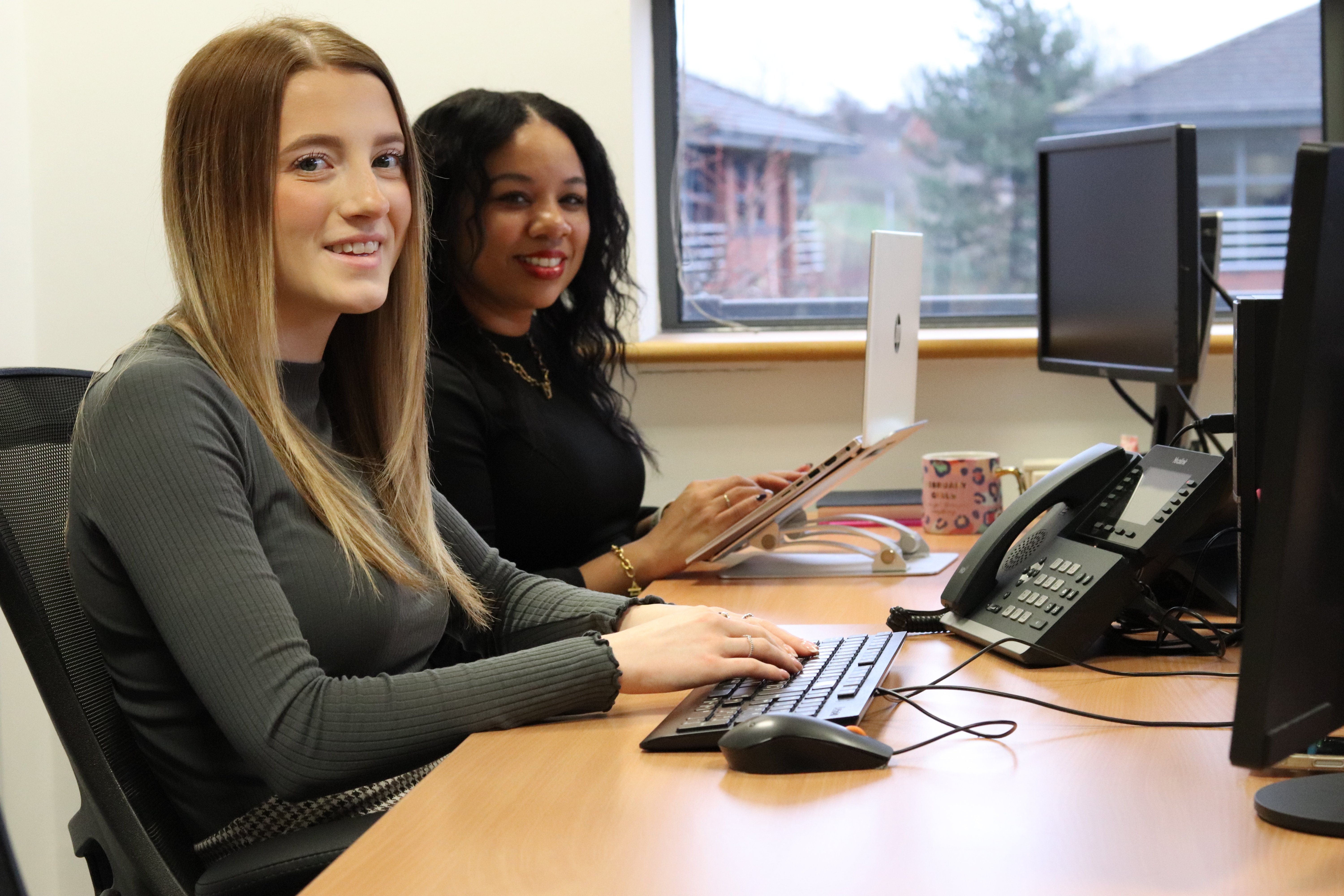 Ellie and Kerry smiling at the camera while typing on their computer keyboards at their desks.