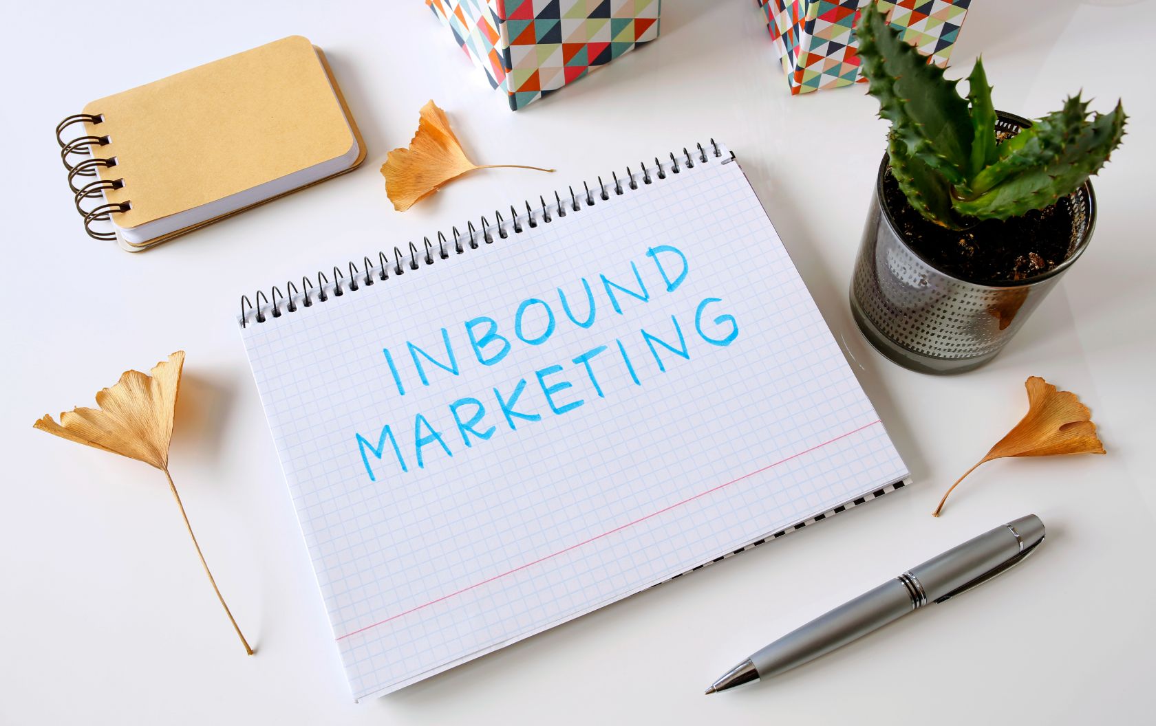 An owner of an SME writing the words Inbound Marketing on a notebook next to a cactus pot, flower, pen, and petals as they start creating their inbound strategy