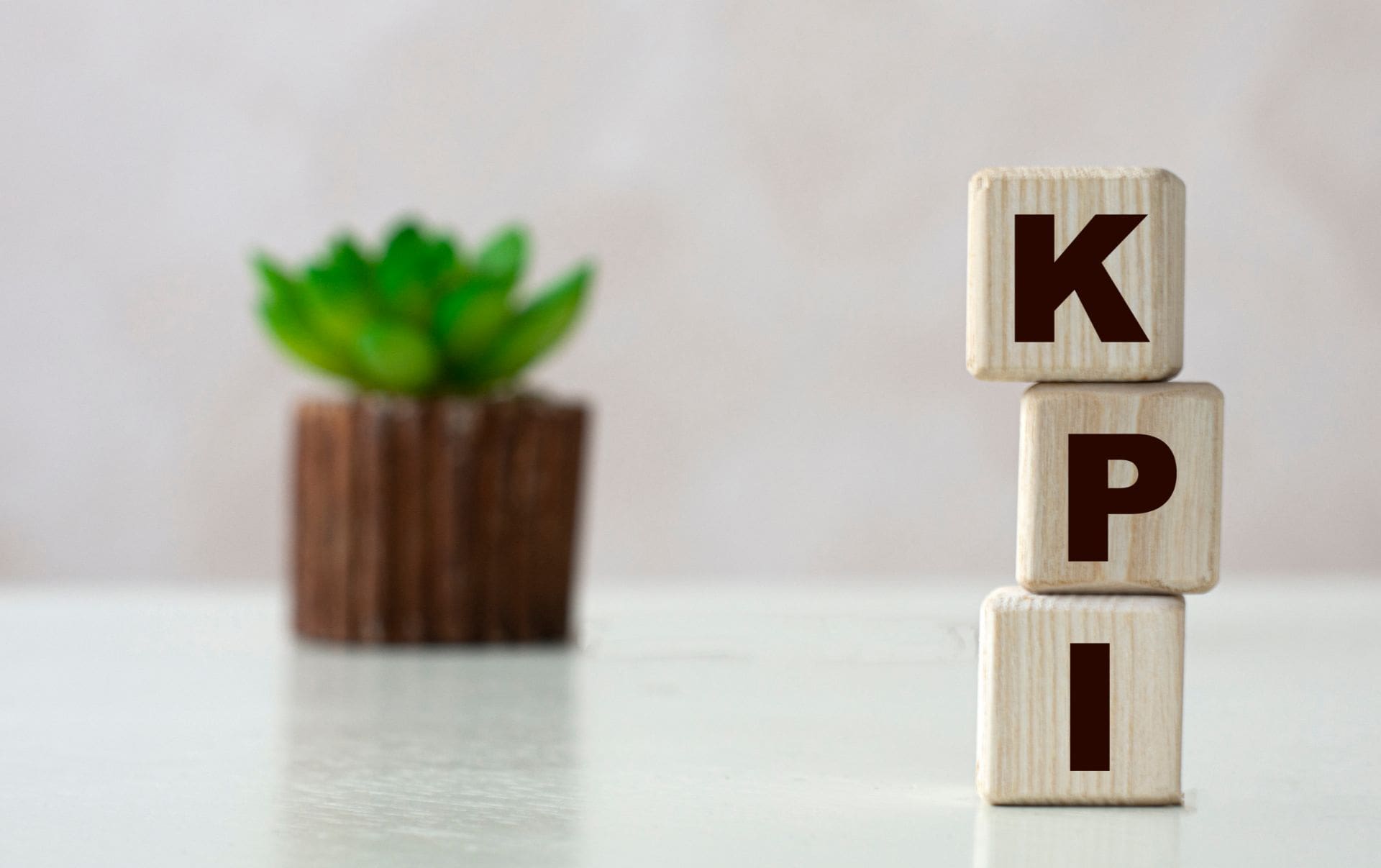 Three wooden blocks that spell out K P I with a succulent in a plant pot behind on the desk of a business owner who is learning how to better monitor their marketing and sales KPIs and metrics. 