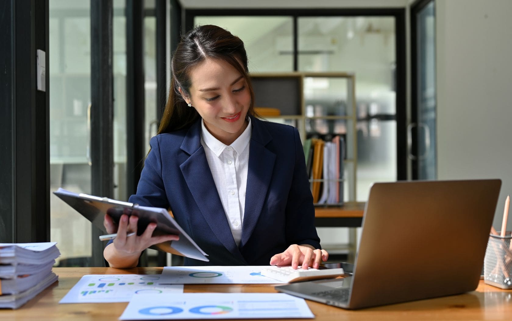 A UK business owner working on her laptop at her desk in an office while reading sales charts from a clip board after her business started following essential sales tips to help it grow.