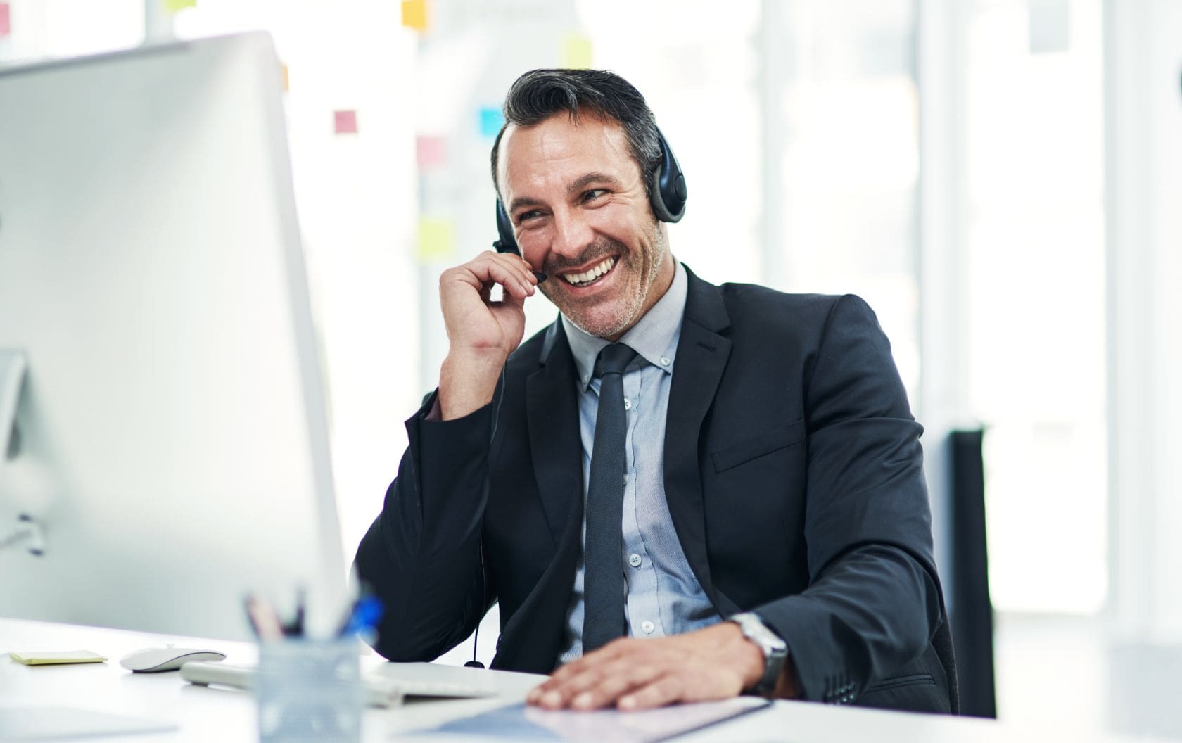 An small business owner at his desk with a desktop before him that he looks at and a headset on where he is talking to a lead about to win a sale masterfully. 
