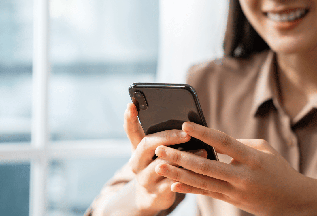 A person holding and using a smartphone. The background includes a window with light coming through it. The person's hands are visible, interacting with the phone's screen.
