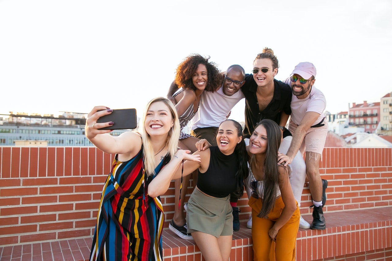Seven millennials huddled together with some sitting on a brick wall as they take a group selfie for social media which businesses can learn from to help grow their engagement 