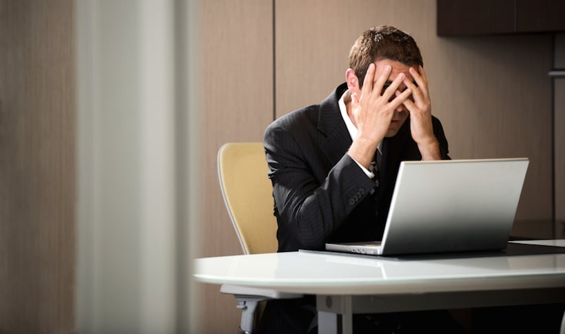 A man seated at a table, sitting in front of his laptop whilst looking stressed due to not knowing enough about marketing.