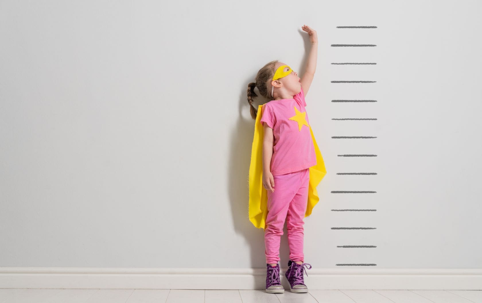 A future business owner as a child in a superhero costume standing on her tip toes trying to grow next to a wall with a height measurement drawn on it which is taller than her.