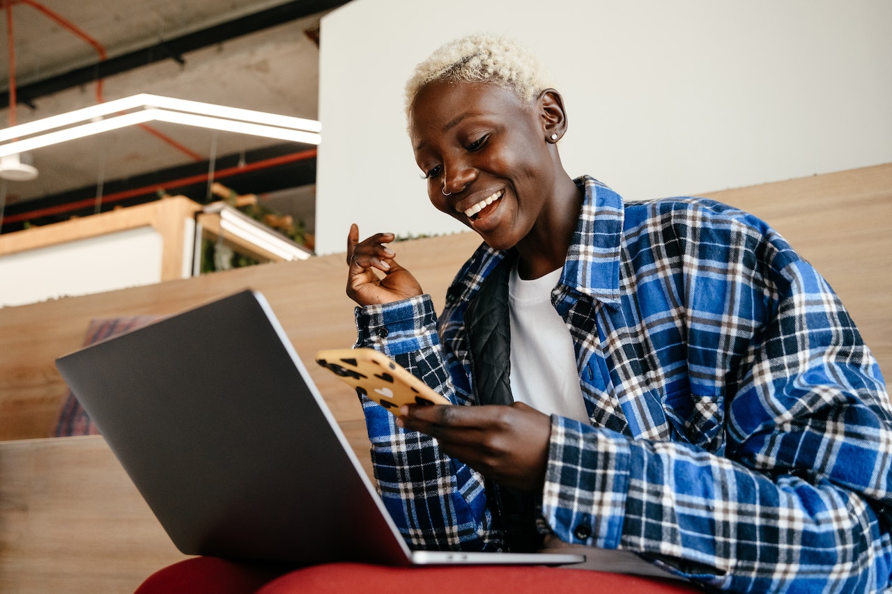 A woman smiling at her phone with her laptop on her lap as she receives sale leads from her small business Chatbot from her website